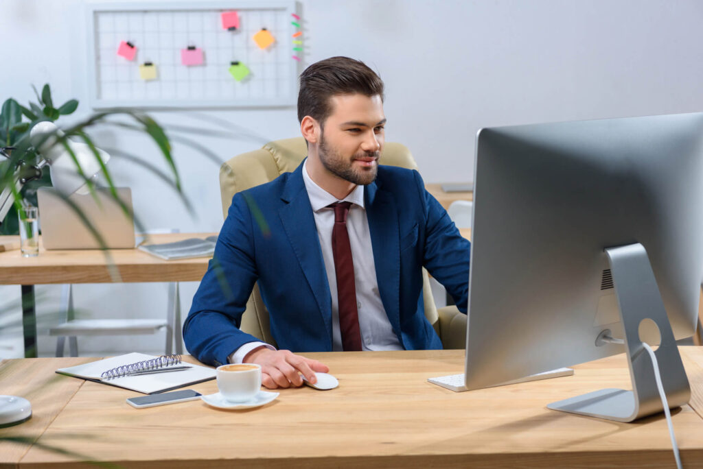 An image of a smiling man (who represents a financial advisor) in a suit sitting at a desk working on a computer .